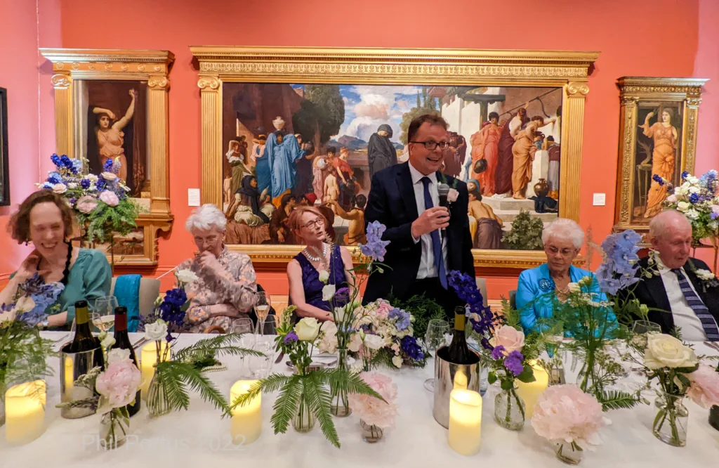 A long table is set for a formal dinner in front of large framed paintings with guests seated and smiling.