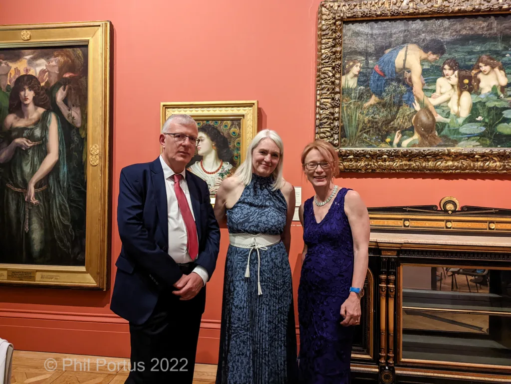 Three people stand between framed paintings on a red wall.