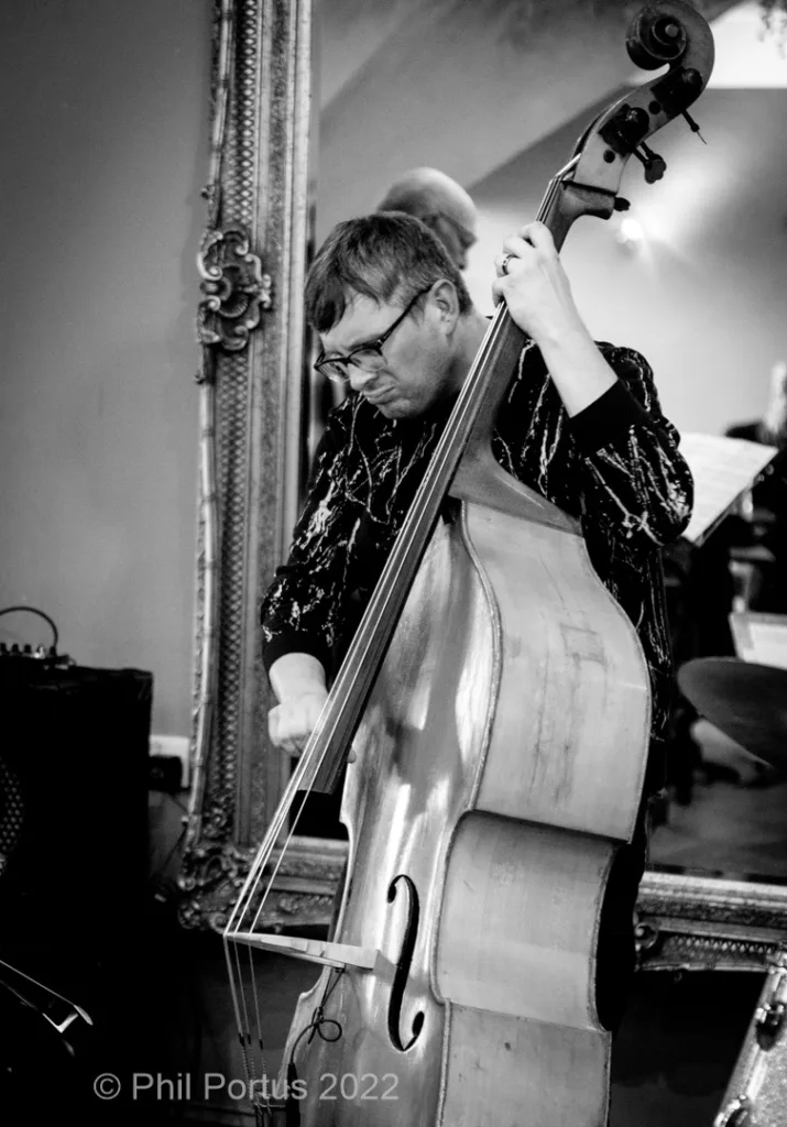 A musician plays a double bass while standing near an ornate mirror in black and white.