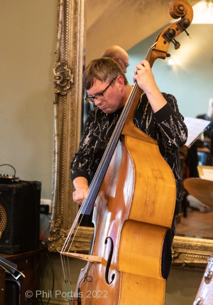 A musician plays a double bass while wearing glasses and a patterned shirt.