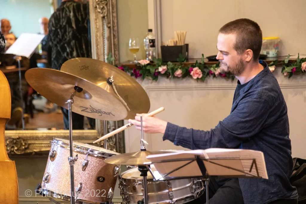 A musician plays drums seated behind a drum kit in an indoor setting with other musicians visible.