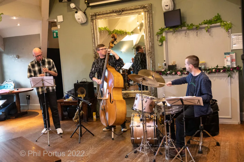 Four musicians perform on a stage with instruments including flute, bass, drums, and keyboard.