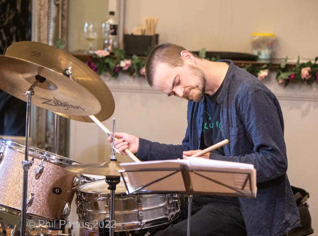 A person with a shaved head plays drums in a dimly lit room decorated with floral arrangements.