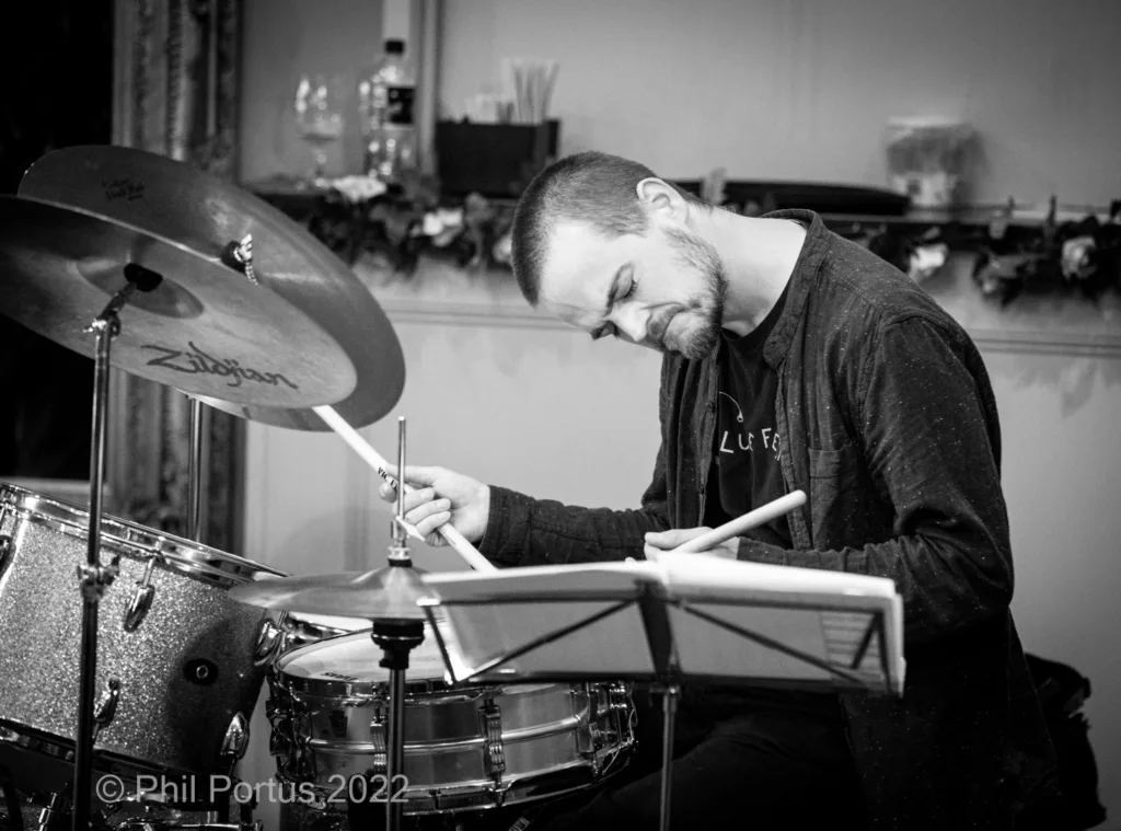 A drummer sits behind a drum kit, striking a cymbal with a drumstick.