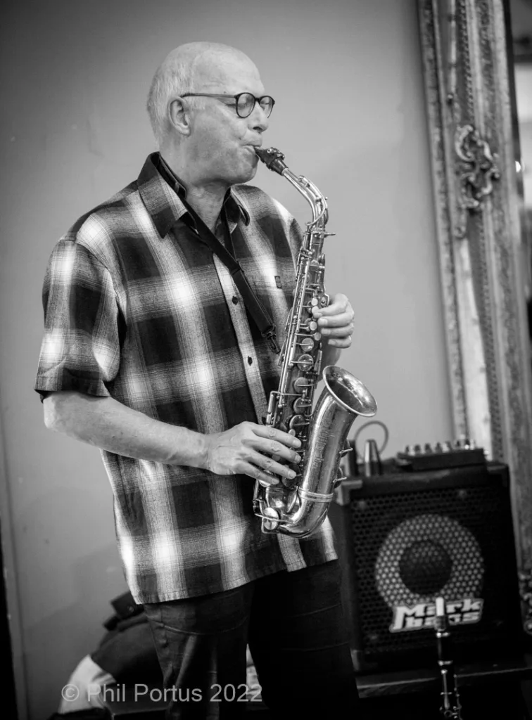 A musician plays a saxophone while wearing glasses and a plaid shirt.