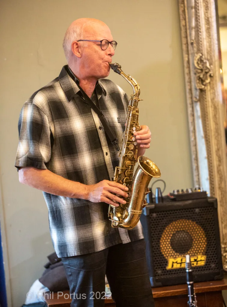 A man with glasses plays a gold saxophone while standing near audio equipment and an ornate mirror.