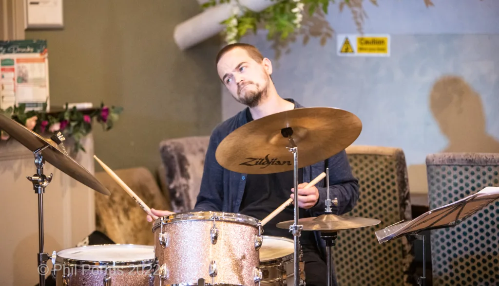 A musician sits behind a drum kit, holding drumsticks and looking toward the camera.