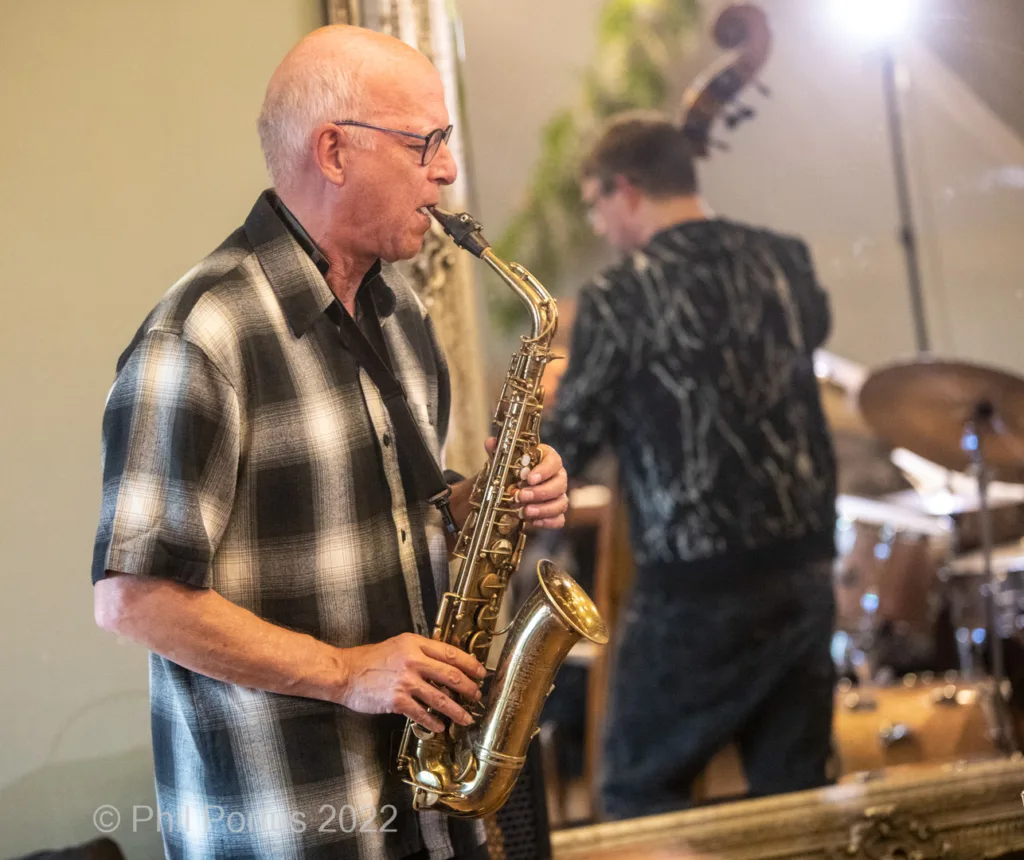 A musician plays a saxophone while another stands behind a double bass in a dimly lit room.