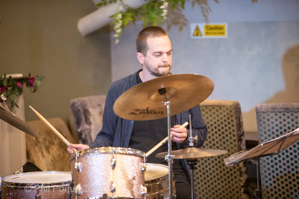 A person with a short beard plays drums in a room with floral decorations and soft lighting.