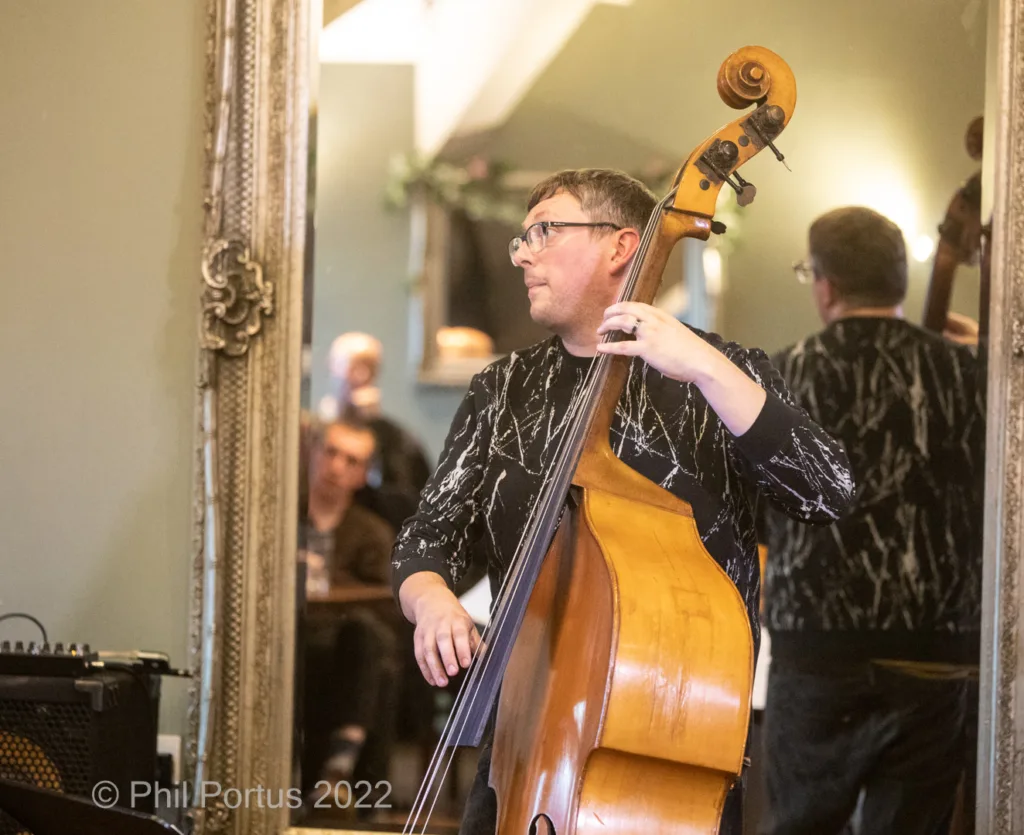 A musician plays a double bass while standing near an ornate mirror and other band members.