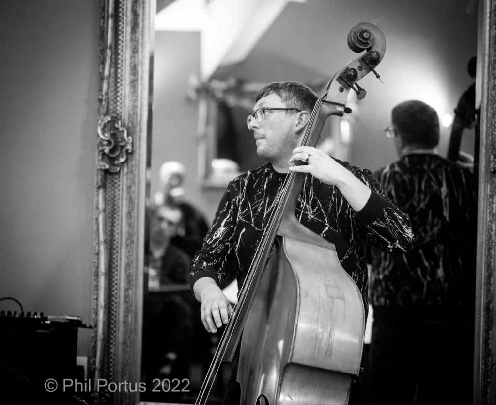 A musician plays a double bass while wearing a patterned shirt and glasses.