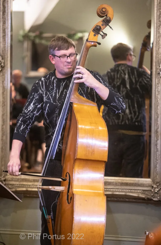 A musician plays a double bass while standing in front of an ornate mirror.