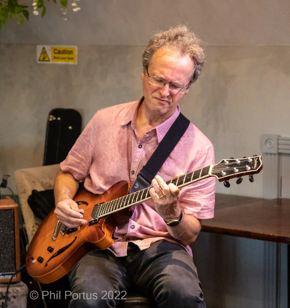 A man with graying hair plays an arched guitar while seated and wearing glasses.