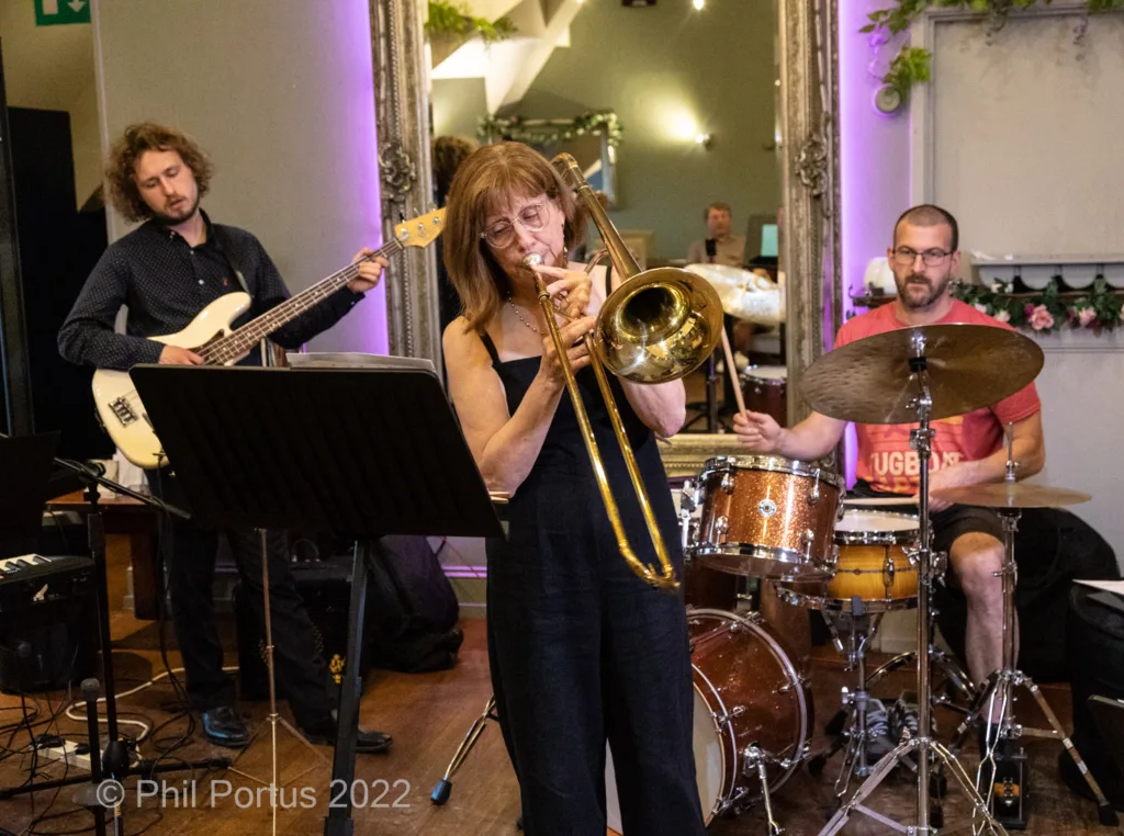 A jazz band performs on a stage with a trombonist in the center and a bassist and drummer visible.