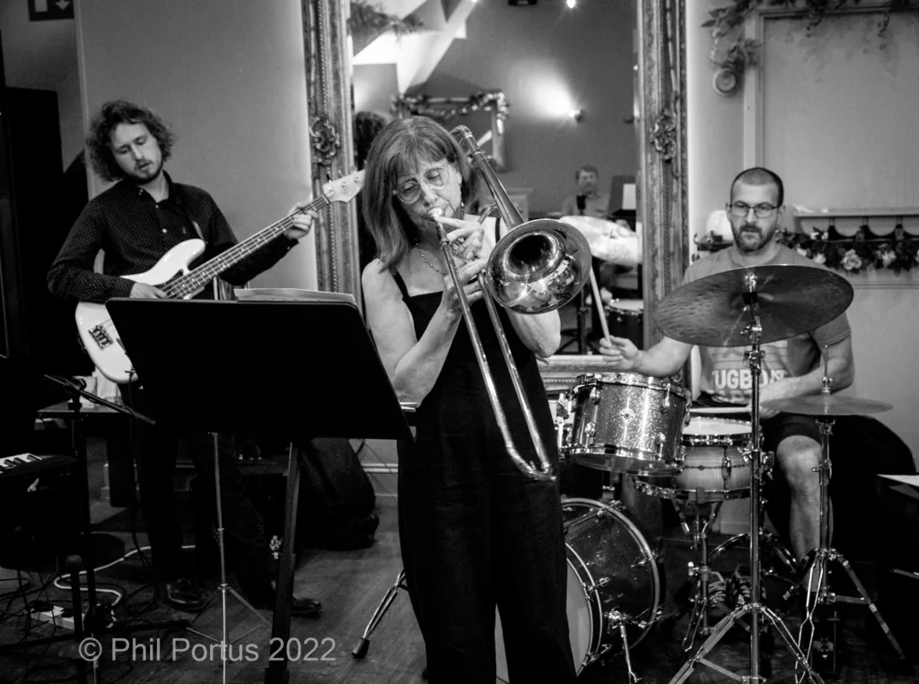 Three musicians perform on a stage with sheet music stands and a large mirror in the background.