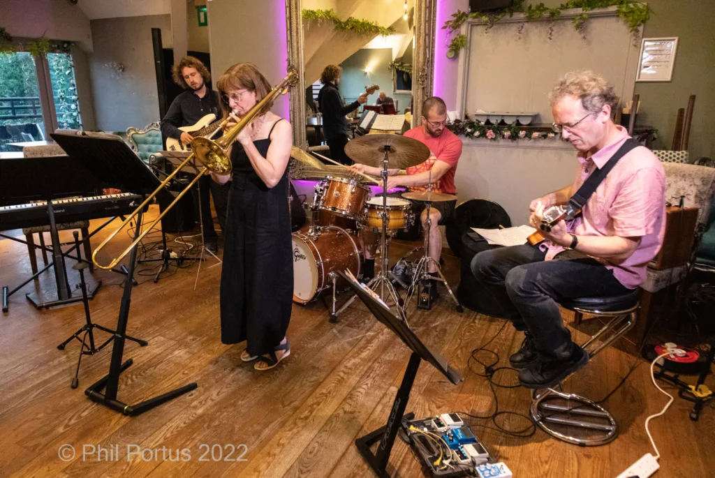 Four musicians perform on a wooden stage in a dimly lit room with decorative greenery.