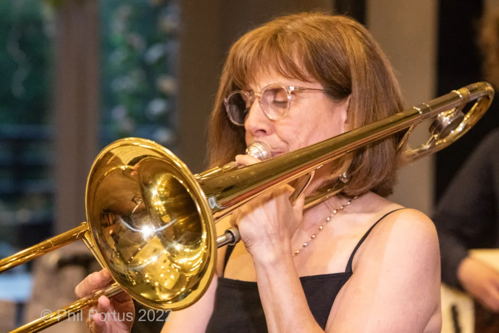 A woman with glasses plays a brass trombone while wearing a black top and silver necklace.