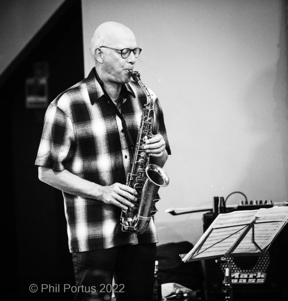 A musician plays a saxophone while wearing glasses and a plaid shirt.
