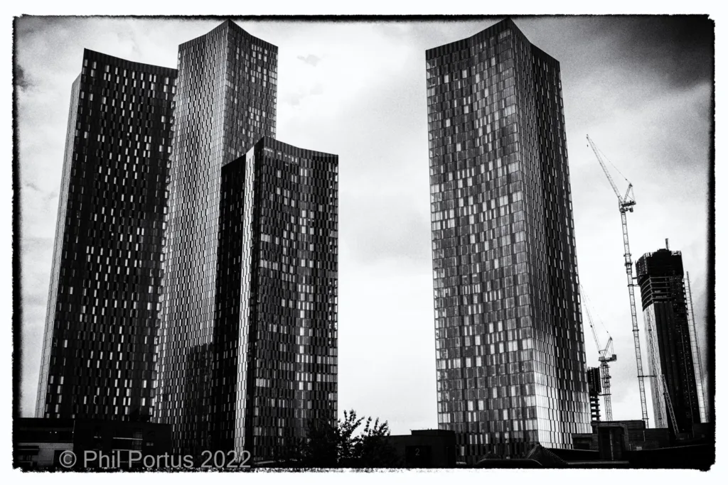 Tall modern buildings with a construction crane are visible against a cloudy sky in black and white.