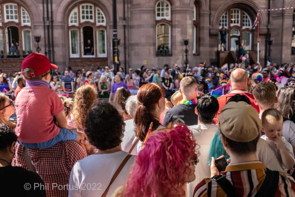 A crowd gathers at an outdoor event with people of various ages and colorful clothing visible.