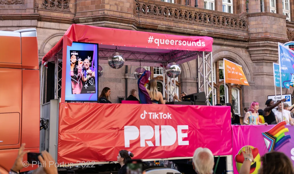 A brightly colored parade float displays a TikTok Pride banner with people waving and dancing nearby.