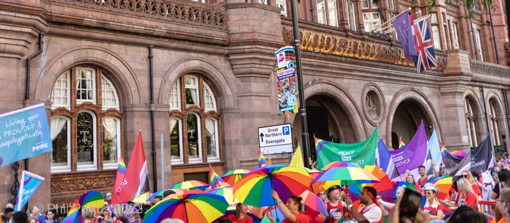 A crowd gathers for a parade holding colorful umbrellas and flags in front of a large stone building.