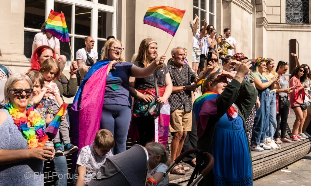 A group of people stand on steps waving rainbow flags at an outdoor event in bright sunlight.