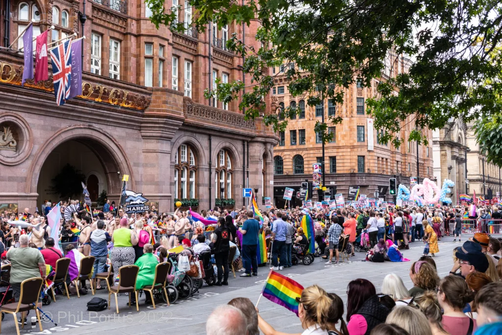 A large crowd gathers on a city street lined with tall brick buildings and flags.
