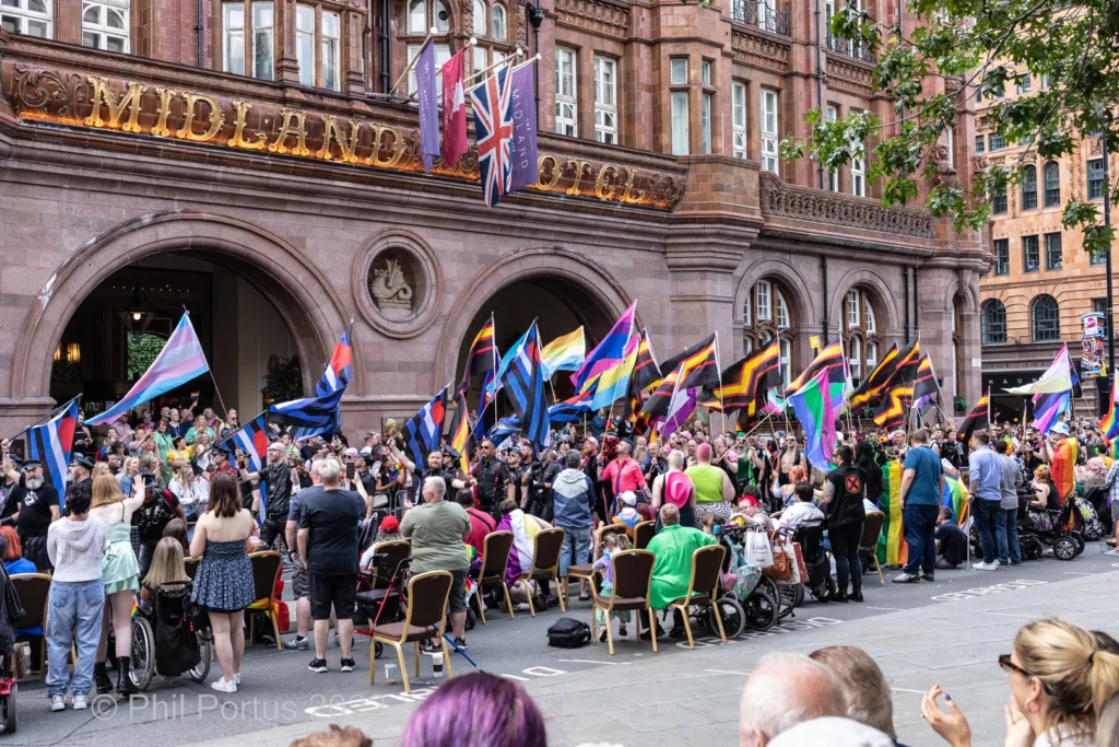 A large crowd gathers outdoors with numerous flags displayed in front of a grand building.