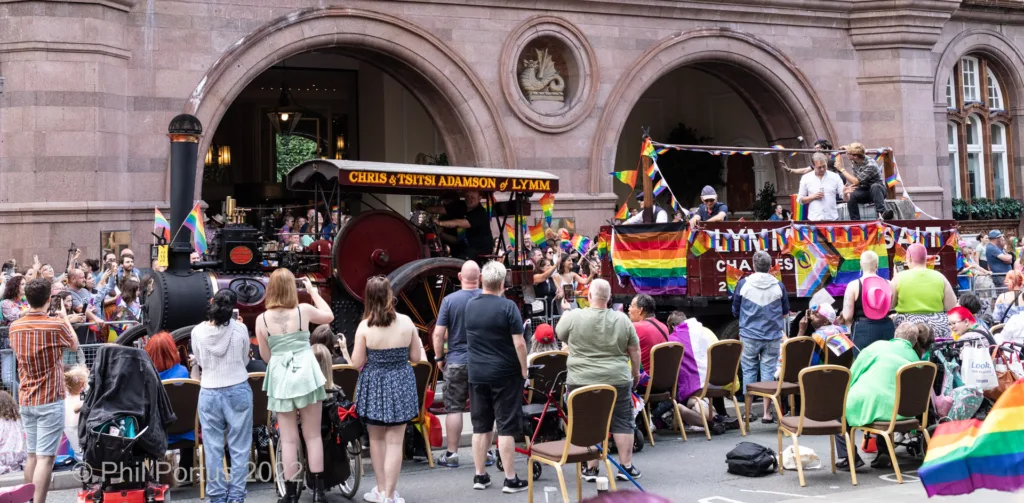 A steam train is decorated with rainbow flags as a crowd gathers on a sunny day.