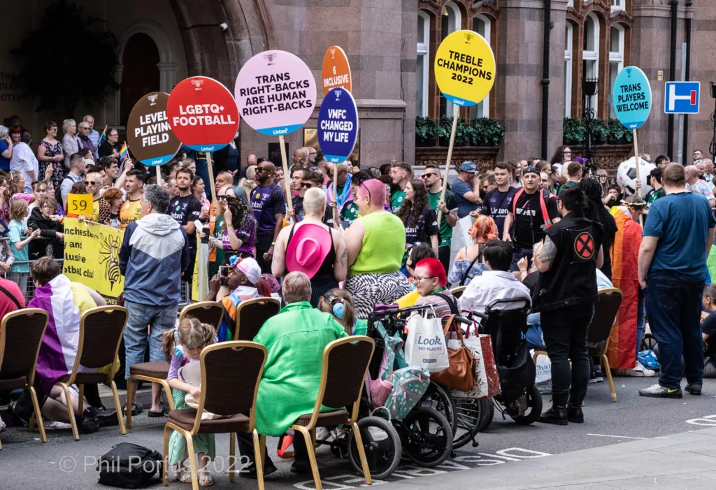 A crowd gathers on a street for an event with signs and chairs visible.