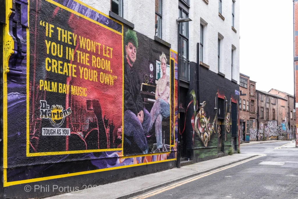 A mural covers a building's exterior wall alongside a street and other buildings.