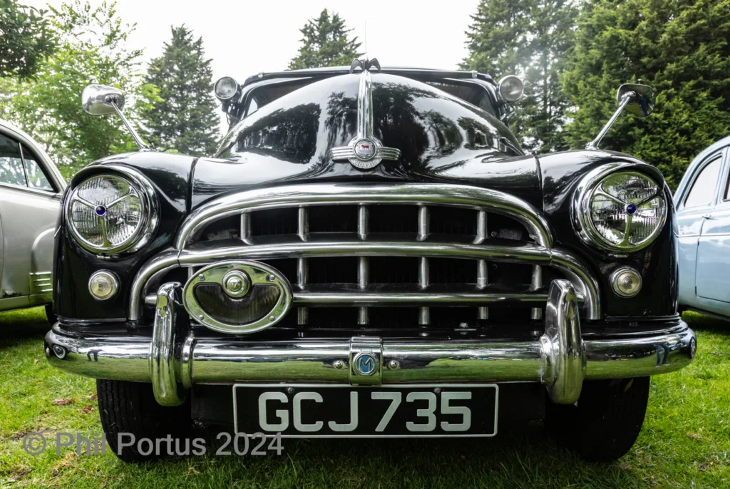A black vintage car with a chrome grill and round headlights sits on green grass.