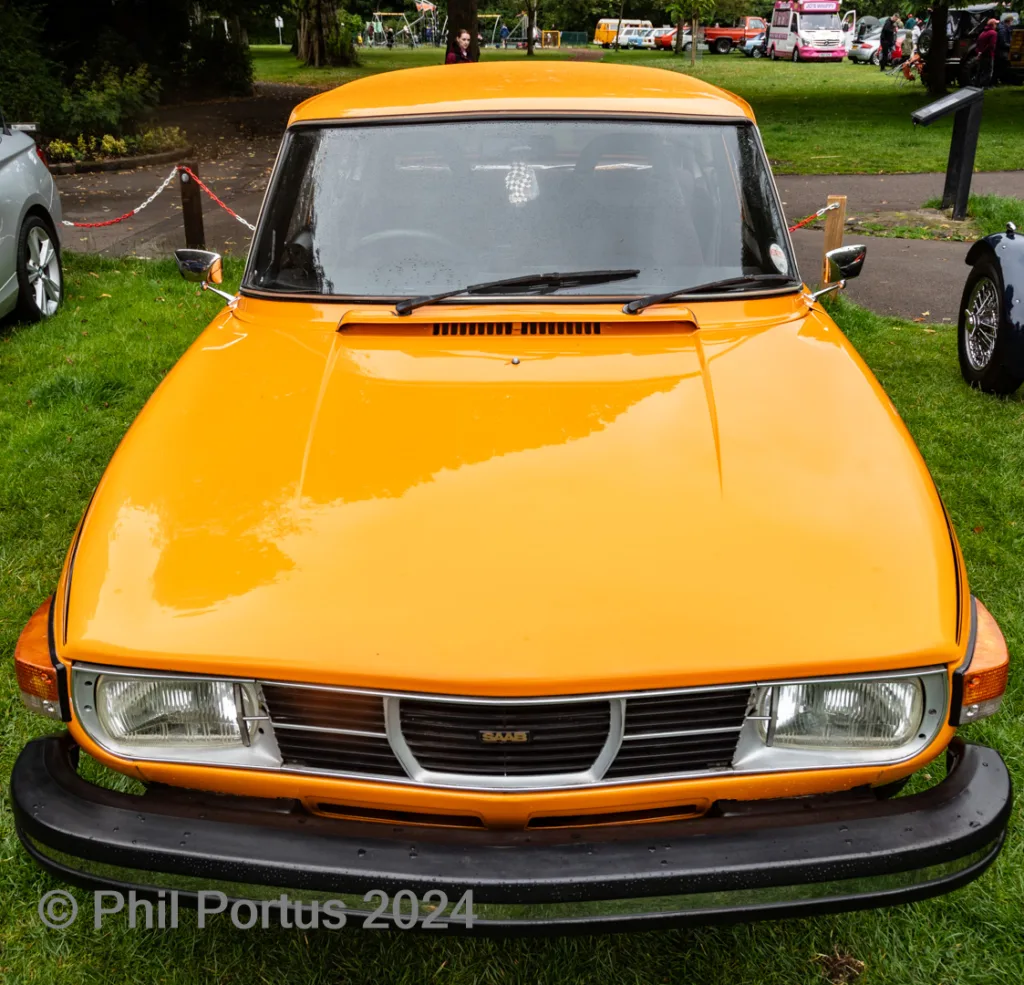 An orange car with a black front spoiler sits on green grass at an outdoor event.