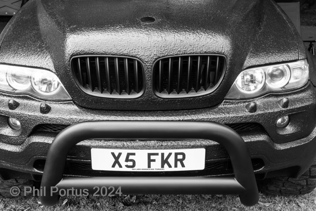 A black and white close-up shows the front of a vehicle with a protective bar and license plate.