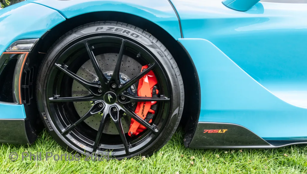 A turquoise sports car's wheel and bright orange brake calipers are visible against a green grassy background.