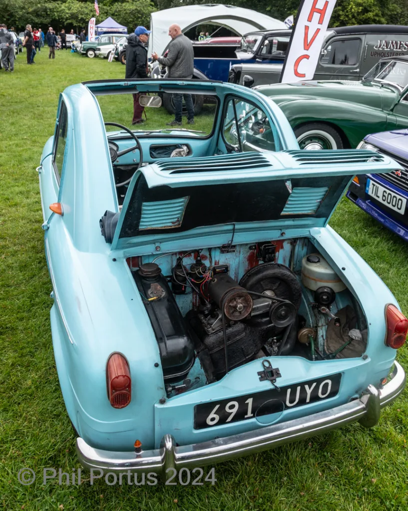 A light blue vintage car with its rear engine exposed sits on a grassy field at an outdoor event.