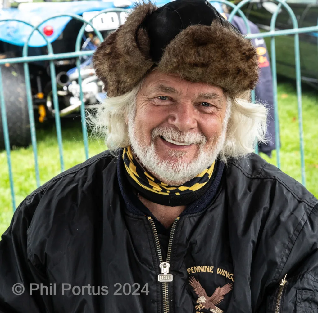 A man with a furry hat and beard smiles while wearing a black jacket with an embroidered patch.