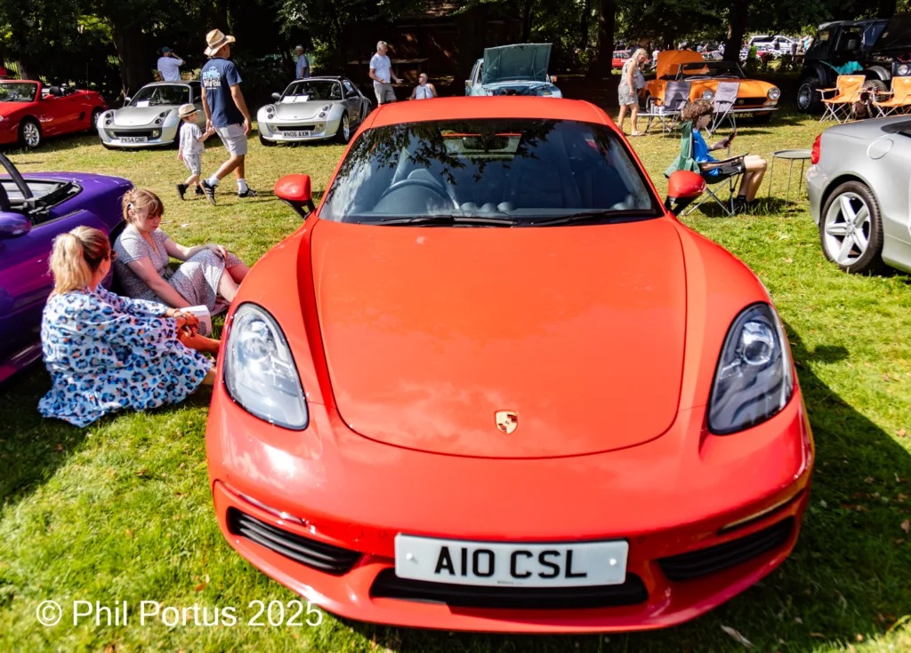 A red sports car sits on a grassy field surrounded by other vehicles and people enjoying the sunshine.