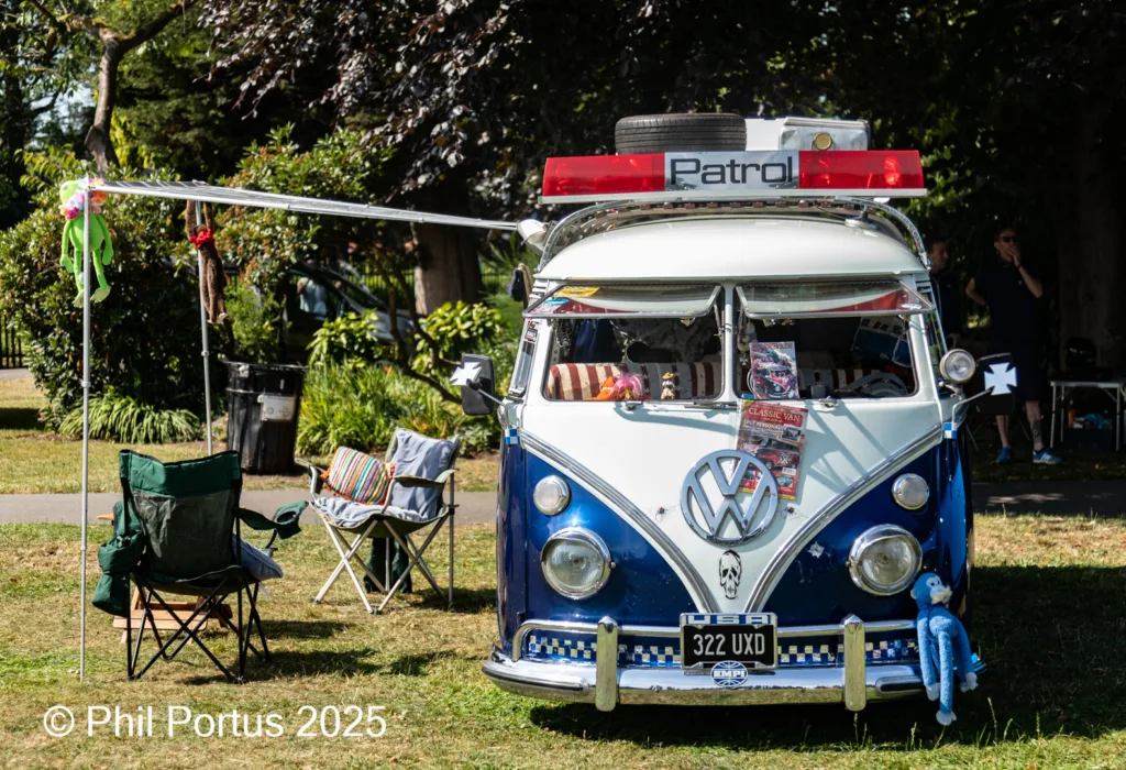 A vintage blue and white Volkswagen van sits on grass with camping chairs nearby.