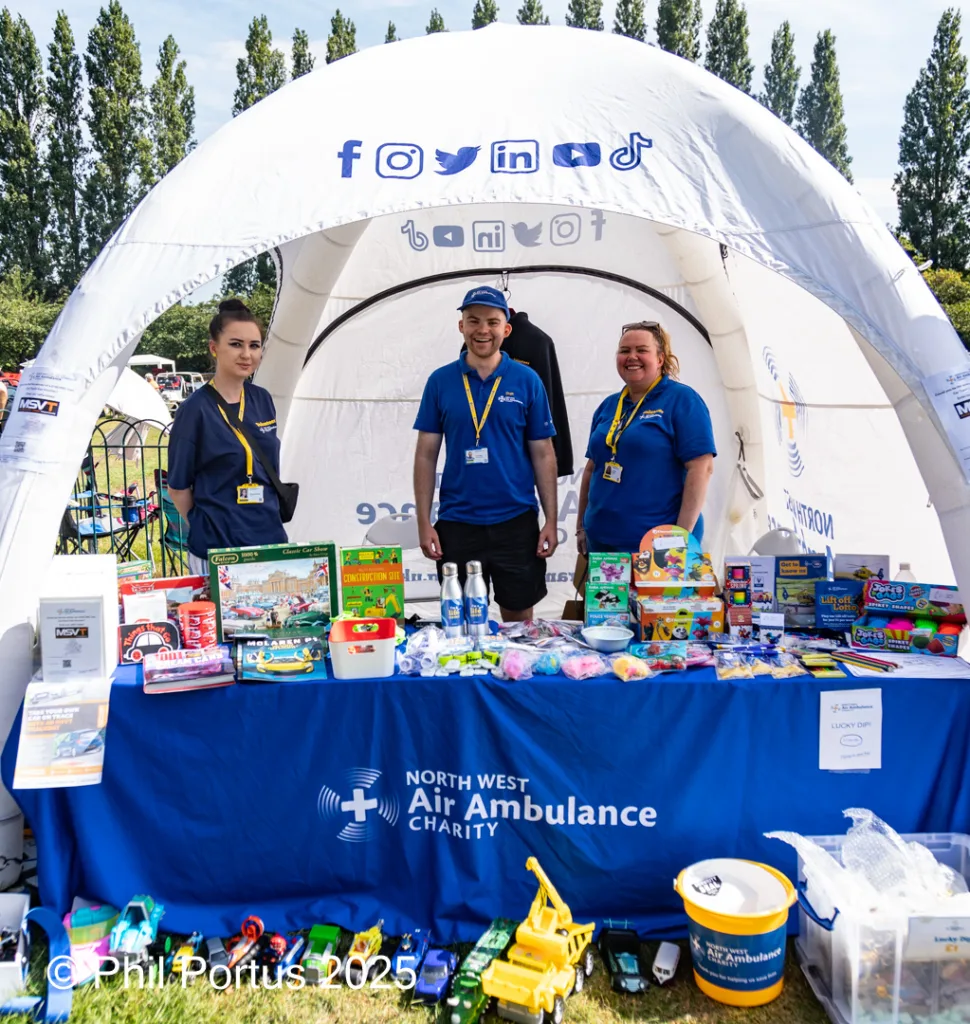 Three people stand behind a table covered in merchandise under a white canopy at an outdoor event.