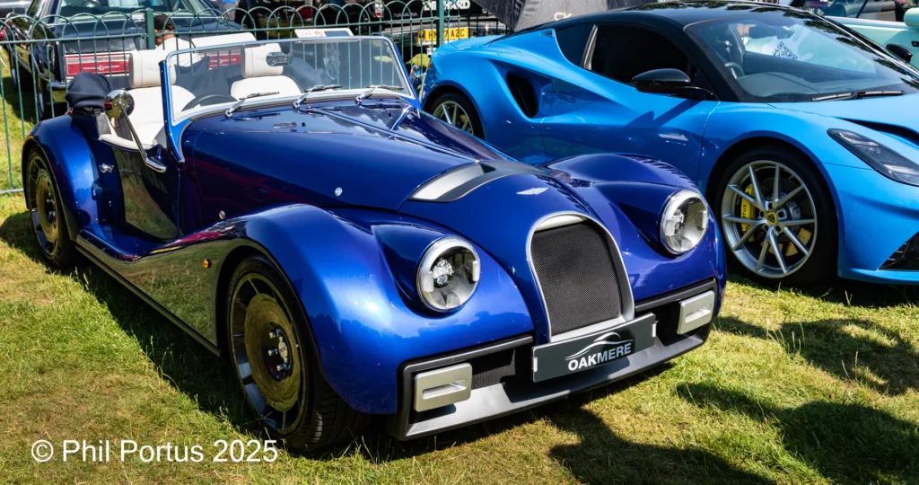 A blue roadster sits on grass next to a dark blue sports car at an outdoor event.