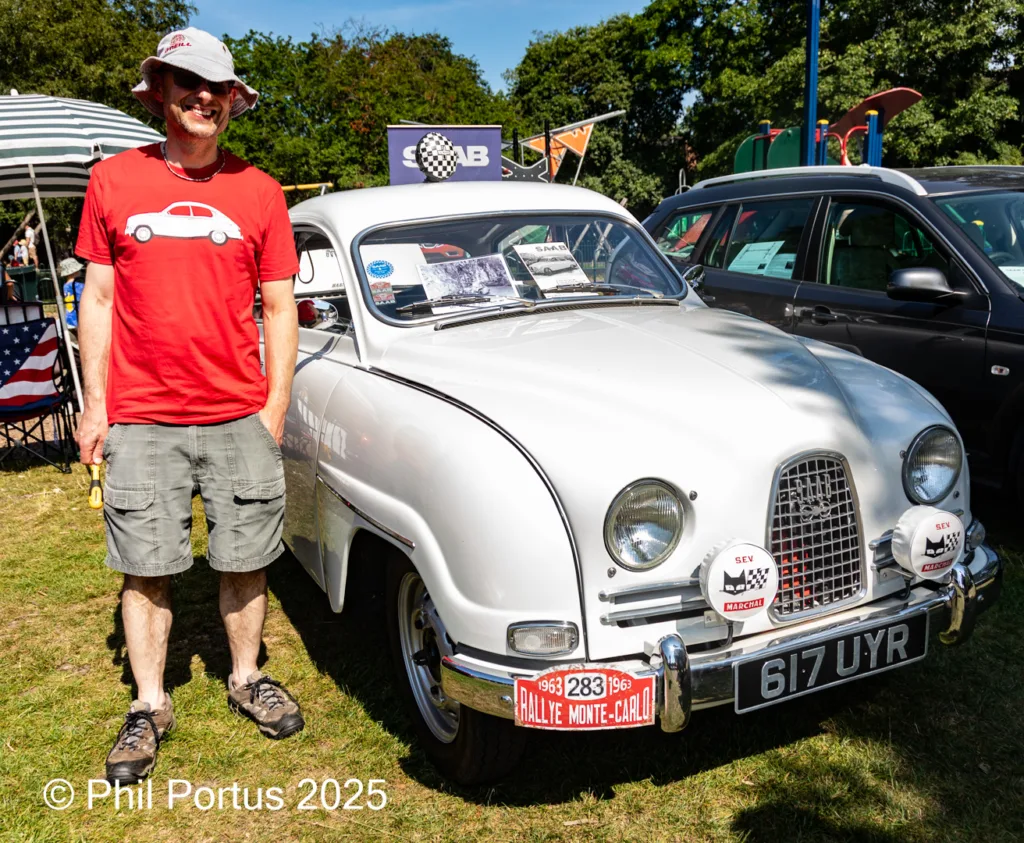 A person stands beside a vintage car at an outdoor event on a sunny day.