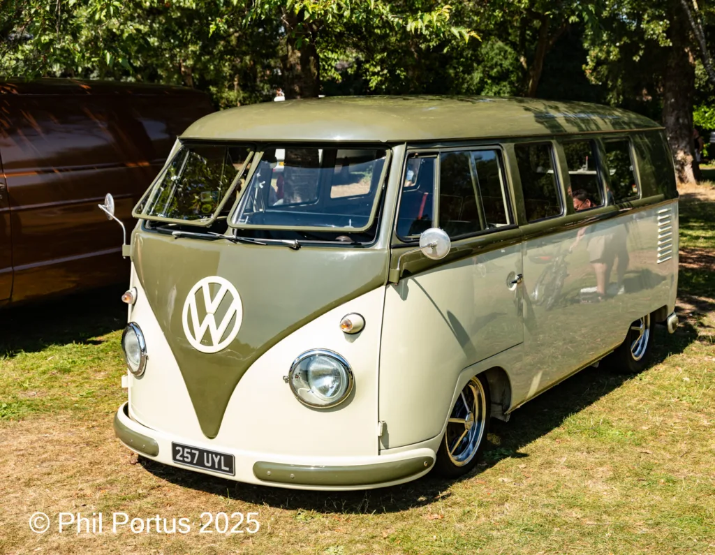 A vintage olive green Volkswagen bus sits on a grassy lawn with trees in the background.