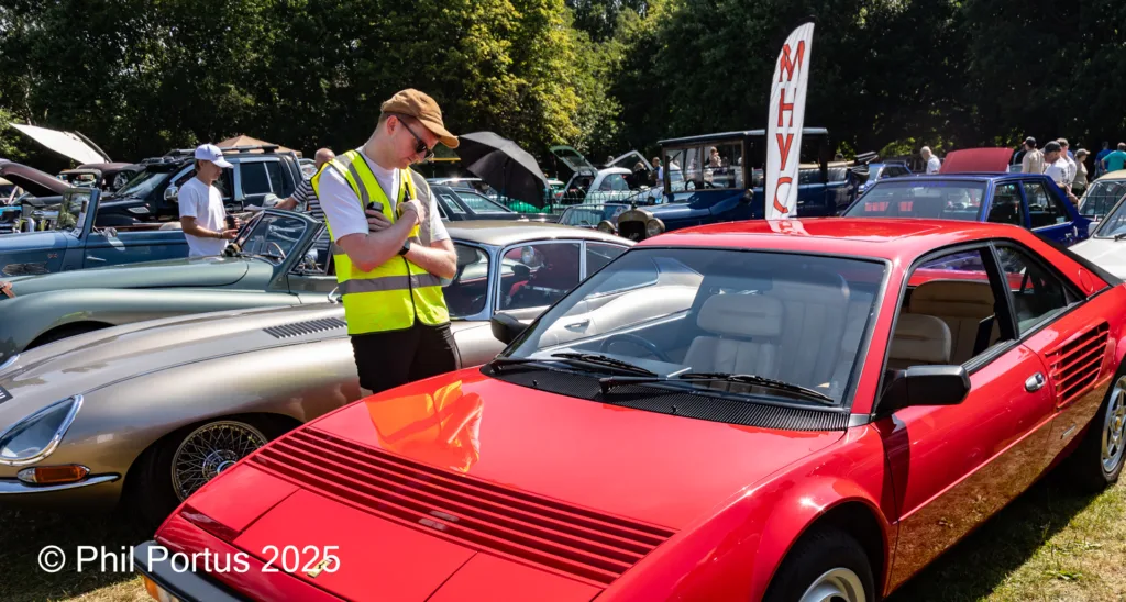 A person wearing a reflective vest looks down at the hood of a red sports car at an outdoor event.
