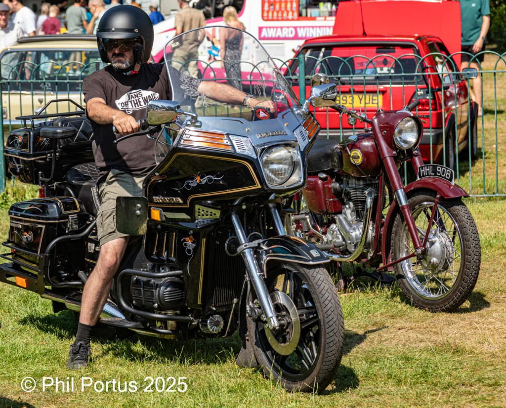 A person sits on a large motorcycle with luggage at an outdoor event alongside other vehicles.