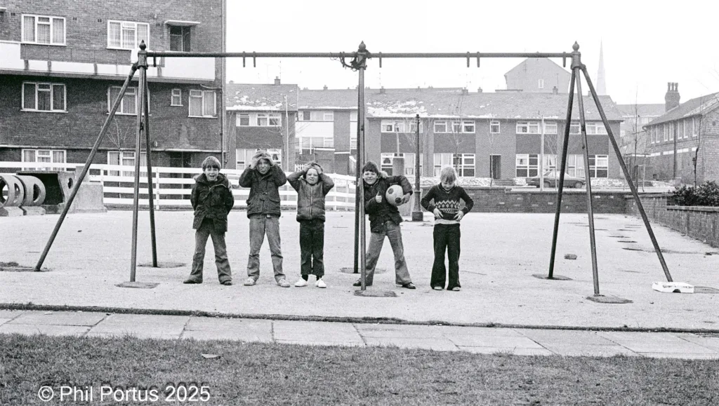 Five children stand in a black and white playground with swings and buildings in the background.