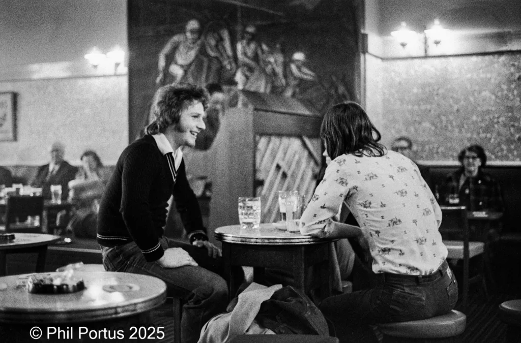 Two people sit at a round table in a dimly lit room with other patrons visible in the background.