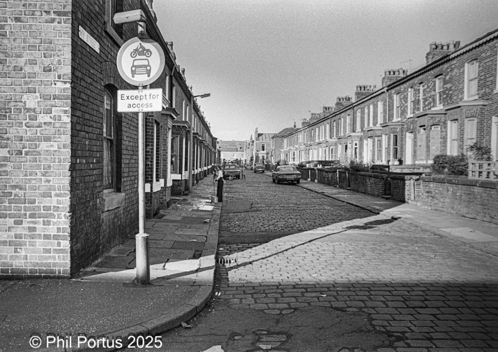 A black and white street scene shows brick buildings lining a road with a traffic sign on the left.