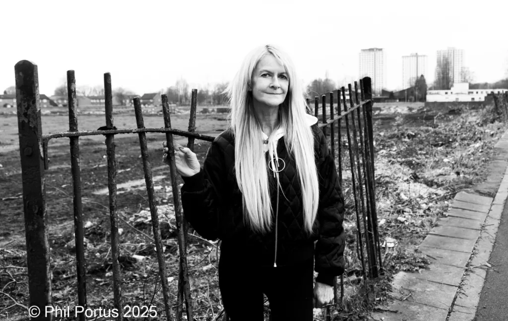 A person with long blonde hair stands near a metal fence in black and white.